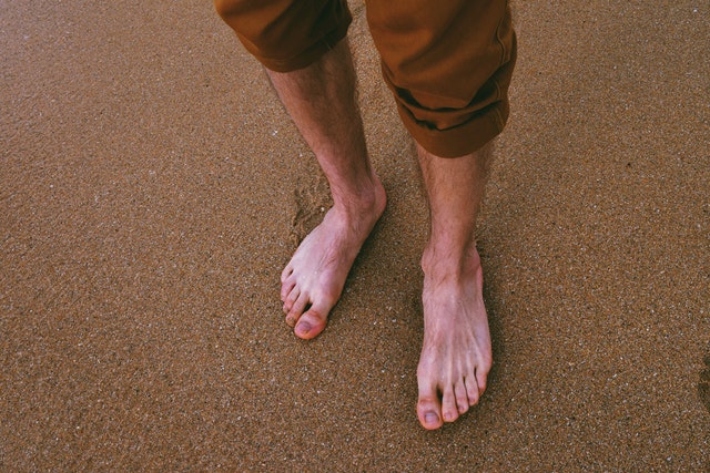 Muž v hnedých krátkych nohaviciach s bosými nohami stojí na piesku